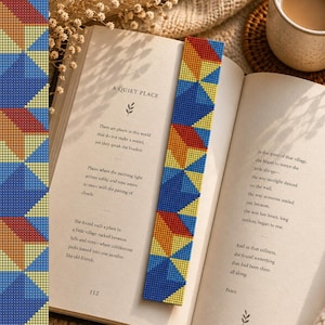 May include: A beaded bookmark with a geometric pattern in blue, yellow, orange, and red is placed in an open book. The book's pages display text about a quiet place. A cup of coffee sits nearby, suggesting a cozy reading setting.