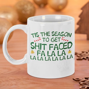 May include: White ceramic mug with the text "TIS THE SEASON TO GET SHIT FACED FA LA LA LA LA LA LA" in green and red. The mug has a curved handle and is set against a festive background with Christmas ornaments.