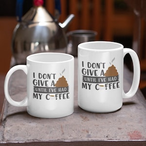 May include: Two white coffee mugs with a brown poop emoji and the text "I don't give a until I've had my c-ffee" on them. The mugs are sitting on a wooden table.