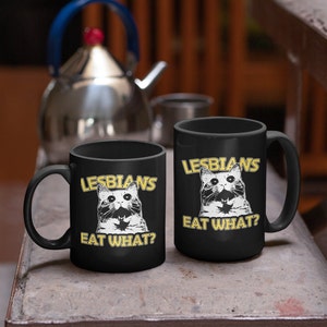 May include: Two black ceramic mugs with white cat illustrations and the text "Lesbians Eat What?" in yellow. The mugs are on a gray surface.