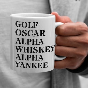 May include: White ceramic mug with the words "GOLF OSCAR ALPHA WHISKEY ALPHA YANKEE" printed in black. The mug is held by a person wearing a gray jacket and white t-shirt.