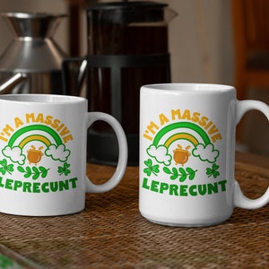 May include: Two white ceramic mugs with the text "I'M A MASSIVE LEPRECUNT" in green and orange. The design includes a rainbow, pot of gold, and shamrocks. The mugs are on a woven surface.