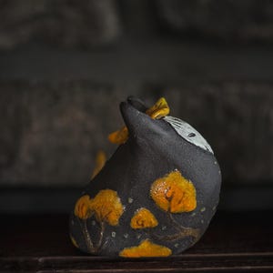 Könnte beinhalten: Eine kleine, dunkelgraue Keramikskulptur in Form eines stilisierten Tieres mit einem spitzen Ohr. Die Oberfläche zeigt handgemalte, leuchtend orangefarbene Baummotive mit strukturierten braunen Stämmen und einen weißen Fleck am oberen Körper.