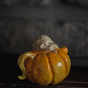 Könnte beinhalten: Eine kleine Keramik-Teekanne in Form eines leuchtend orangefarbenen Kürbisses mit einer strukturierten, hellbraunen Igel-Figur, die oben herausragt. Die Teekanne hat einen geschwungenen Henkel auf der linken Seite und einen kleinen Ausguss auf der rechten Seite mit glänzender Oberfläche.