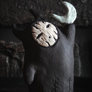 Peut inclure: Une figurine en céramique gris anthracite à la surface texturée. Le personnage présente un visage rond couleur crème avec des lignes gravées, deux petites cornes et un bras levé tenant un croissant de lune vert pâle brillant.