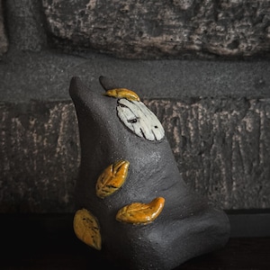 Peut inclure: Une sculpture en céramique gris foncé avec une finition mate et lisse. L'objet présente un visage blanc de forme ovale avec des marques noires simples et trois petites feuilles jaunes brillantes fixées sur le côté. La forme est arrondie.