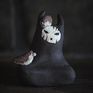 Op de afbeelding: Een donker antracietkleurig keramisch beeldhouwwerk met een centraal wit maskerachtig gezicht met geëtste details. Twee kleine bruin-witte vogels zitten op het figuur, één op het hoofd en één aan de zijkant, met een gladde basis.