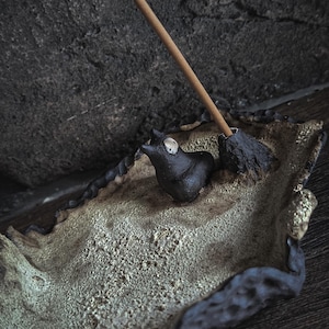 Può includere: Un porta incenso in ceramica dalla consistenza irregolare, con un bordo scuro e grezzo e un interno color sabbia chiaro. Una piccola figura animale scura è posizionata sulla base accanto a un supporto conico scuro con un bastoncino.