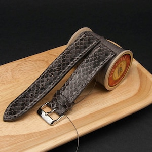 Op de afbeelding: Een grijs leren horlogebandje met een slangenhuidpatroon. Het bandje wordt getoond op een houten oppervlak met een spoel draad.