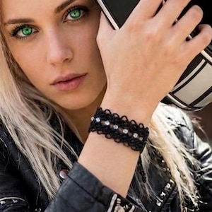 May include: A black lace bracelet featuring small silver-toned accents and clear beads. The bracelet is worn on a wrist, paired with a black leather jacket with silver zippers and studs, and a black motorcycle helmet with white stripes.