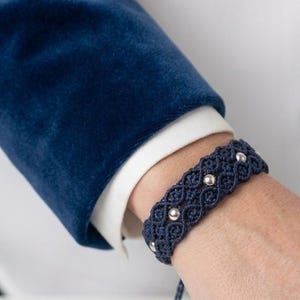 May include: A dark blue macrame bracelet featuring a woven cord pattern and small, round, polished silver-toned metal beads embedded along the center. The bracelet has adjustable cord ends and a secure, knotted design that wraps around the wrist.