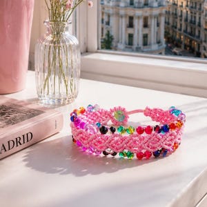 May include: A pink macrame bracelet featuring a woven pattern with integrated multi-colored beads in shades of red, blue, green, and yellow. The bracelet has a circular pink cord closure and is positioned on a white surface near a book.