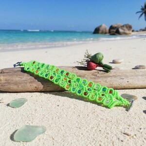 Puede incluir: Una pulsera de macramé de color verde lima brillante con un patrón de cordón tejido y cuentas translúcidas incrustadas en tonos rojo, naranja, amarillo y verde. La pulsera se asegura con un cierre de palanca de metal y descansa sobre madera flotante.