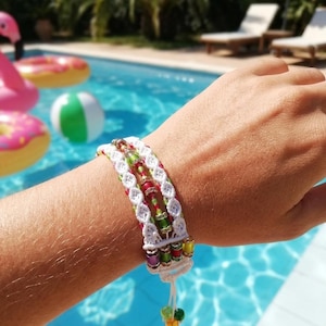 Puede incluir: Una pulsera hecha a mano con un diseño de macramé blanco con cuentas de colores en rojo, verde y amarillo. La pulsera está adornada con una variedad de cuentas y una borla colgante con cuentas de colores. La pulsera se lleva en una muñeca.
