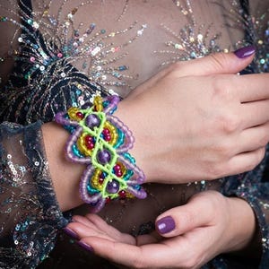 May include: A wide macrame bracelet featuring a bright neon green cord woven into a complex pattern. The design incorporates translucent purple, yellow, and teal beads, along with larger round purple stones arranged in a central vertical line.