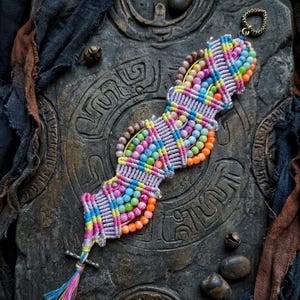 Puede incluir: Una colorida pulsera de macramé con una variedad de cuentas en tonos rosa, naranja, amarillo, verde y azul. La pulsera tiene una borla y un cierre de metal. La pulsera se muestra sobre una superficie oscura y texturizada.