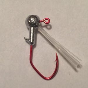 May include: A silver jig head with a red hook and a clear plastic tube. The hook is curved and has a sharp point. The jig head has a loop for attaching to fishing line. Fishing tackle.