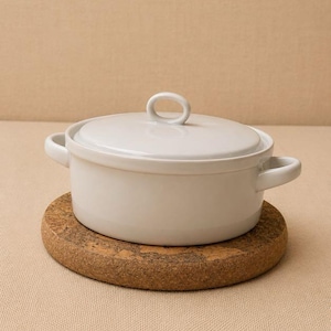 May include: A white ceramic casserole dish with a matching lid and two side handles. The round pot features a smooth, glossy finish and a central loop handle on the lid. It is placed on a circular, textured brown cork trivet for heat protection.