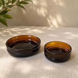May include: Two circular bowls made from dark amber glass. The bowls feature a smooth, rounded rim and a slightly curved base, with one bowl noticeably larger in diameter than the other. The glass is translucent, showing a deep brown hue.