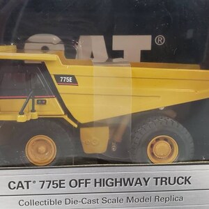 Op de afbeelding: Een geel die-cast model van een CAT 775E off-highway truck. De truck staat op een zwarte en gele doos met de tekst "CAT 775E OFF HIGHWAY TRUCK" en "Collectible Die-Cast Scale Model Replica".