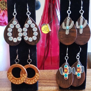 May include: Assortment of wooden earrings with various designs.  The earrings feature a floral design, a cactus design, and a wreath design.  The earrings are displayed on black stands.  "Every journey starts with one step."