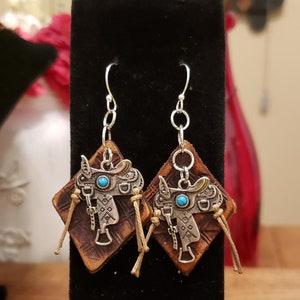 May include: A pair of dangle earrings with a brown leather diamond-shaped charm featuring a silver saddle with a turquoise stone. The earrings have silver hooks and chains.