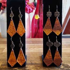 May include: Three pairs of dangle earrings displayed on black velvet stands. The earrings are made of leather and have a diamond shape. The top of each earring features a different metal charm.