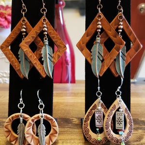 May include: Three pairs of leather earrings with different designs. The first pair is a diamond shape with a turquoise feather dangle. The second pair is a round shape with a turquoise feather dangle. The third pair is a teardrop shape with a silver charm and turquoise beads.