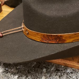 May include: A black cowboy hat with a brown leather band and a decorative design. The band has a small metal stud. The hat has a brown leather cord with wooden beads attached.