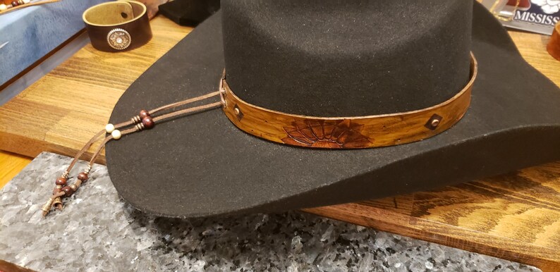 May include: A black cowboy hat with a brown leather band and a decorative design. The band has a small metal stud. The hat has a brown leather cord with wooden beads attached.