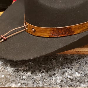 May include: A black felt cowboy hat with a brown leather band featuring a floral design. The hat has a brown leather chin strap with beads.