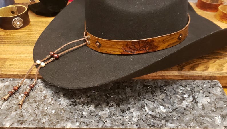 May include: A black felt cowboy hat with a brown leather band featuring a floral design. The hat has a brown leather chin strap with beads.