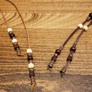 May include: Two brown leather cords with brown and white beads and metal accents. The cords are tied at the ends.
