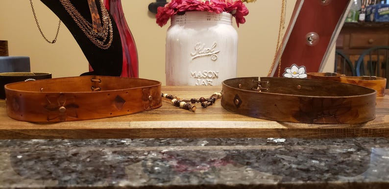 May include: Two brown leather belts with decorative floral designs. The belts are on a wooden surface.