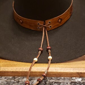 May include: A black cowboy hat with a brown leather band and a beaded leather chin strap. The hat has a wide brim and a tall crown.