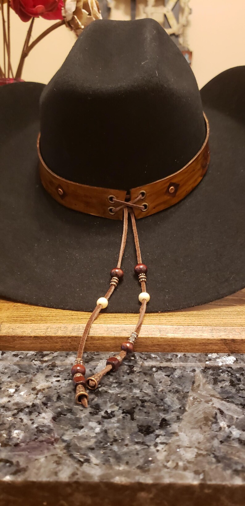 May include: A black cowboy hat with a brown leather band and a beaded leather chin strap. The hat has a wide brim and a tall crown.