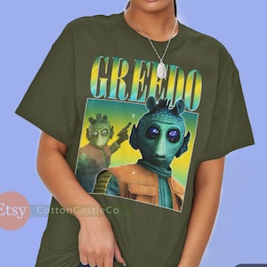 May include: A green t-shirt with a graphic print of a character from the Star Wars movie franchise. The character is a green alien with large eyes and a yellow and blue background. The text "GREEDO" is printed in yellow and blue letters on the shirt.