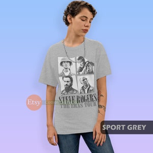 May include: A grey t-shirt with a black and white image of Steve Rogers from the Marvel Cinematic Universe. The image shows Steve Rogers in four different stages of his life, from his childhood to his time as Captain America. The text "Steve Rogers The Eras Tour" is printed below the image.