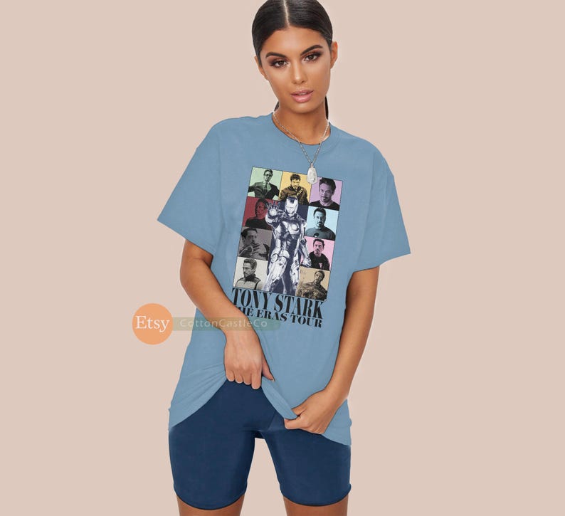May include: Light blue t-shirt featuring a colourful graphic of Tony Stark from the Marvel Cinematic Universe. The graphic depicts different versions of Tony Stark throughout the films. The text "Tony Stark The Eras Tour" is printed below the graphic.