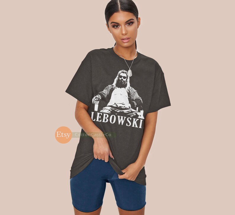 Peut inclure: Un t-shirt gris fonc&eacute; avec un motif blanc d'un homme assis sur un tr&ocirc;ne tenant un verre. Le texte "LEBOWSKI" est imprim&eacute; sous le motif.