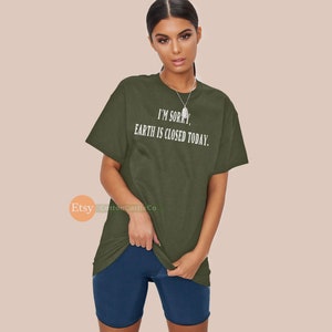 May include: Olive green t-shirt with the text "I'M SORRY, EARTH IS CLOSED TODAY." The shirt is a relaxed fit and has short sleeves. The model is also wearing navy blue shorts.