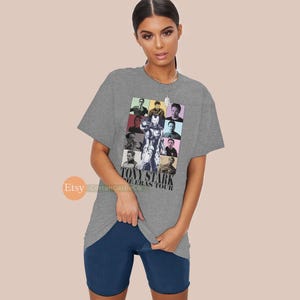 May include: Grey t-shirt featuring a collage of images of Tony Stark from the Iron Man films. The text "Tony Stark The Eras Tour" is printed on the shirt.
