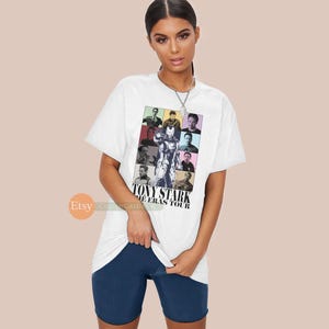 May include: White t-shirt featuring a colourful graphic of Tony Stark from the Marvel Cinematic Universe. The graphic is a collage of different images of Tony Stark throughout his appearances in the Marvel films. The text "Tony Stark The Eras Tour" is printed below the graphic.
