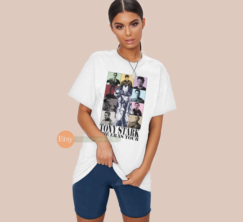 May include: White t-shirt featuring a colourful graphic of Tony Stark from the Marvel Cinematic Universe. The graphic is a collage of different images of Tony Stark throughout his appearances in the Marvel films. The text "Tony Stark The Eras Tour" is printed below the graphic.