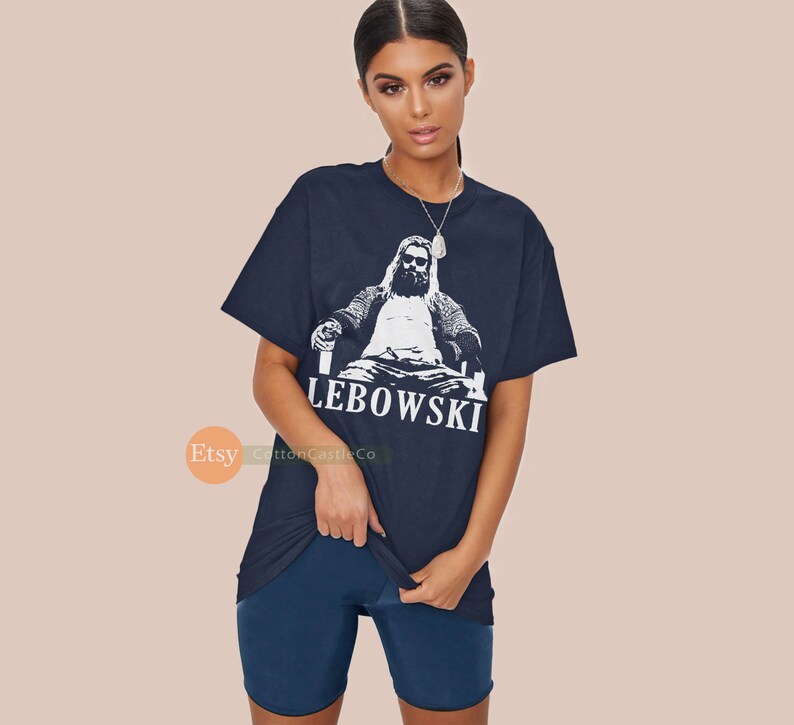 Peut inclure: Un t-shirt bleu marine avec un motif blanc d'un homme assis sur un tr&ocirc;ne avec une boisson &agrave; la main. Le texte "The Lebowski" est imprim&eacute; sous le motif.