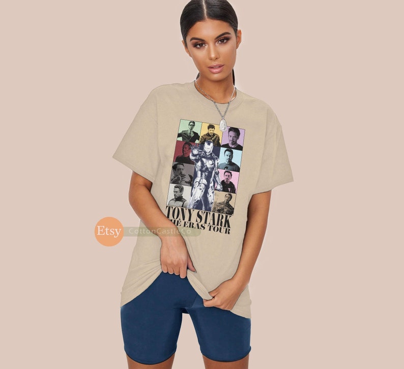 May include: Beige t-shirt featuring a colourful collage of images of Tony Stark from the Iron Man films. The text "Tony Stark The Eras Tour" is printed on the shirt.