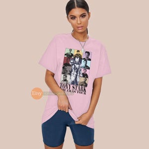 May include: Pink t-shirt featuring a collage of images of Tony Stark from various films. The text "TONY STARK THE ERAS TOUR" is printed on the shirt.