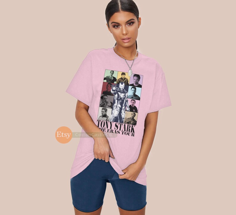 May include: Pink t-shirt featuring a collage of images of Tony Stark from various films. The text "TONY STARK THE ERAS TOUR" is printed on the shirt.