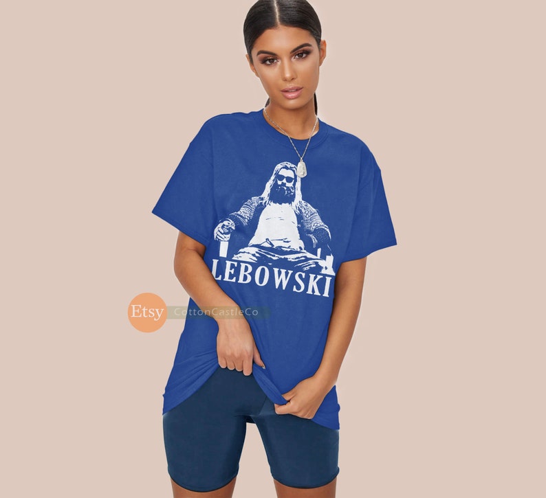 Peut inclure: Un t-shirt bleu avec un motif blanc d'un homme assis sur un tr&ocirc;ne avec une boisson &agrave; la main. Le texte "LEBOWSKI" est imprim&eacute; sous le motif.