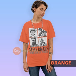 May include: Orange t-shirt with a graphic of Steve Rogers from the Marvel Cinematic Universe. The graphic features four images of Steve Rogers from different points in his life, including his time as a soldier in World War II and as Captain America. The text "Steve Rogers The Eras Tour" is printed below the images.